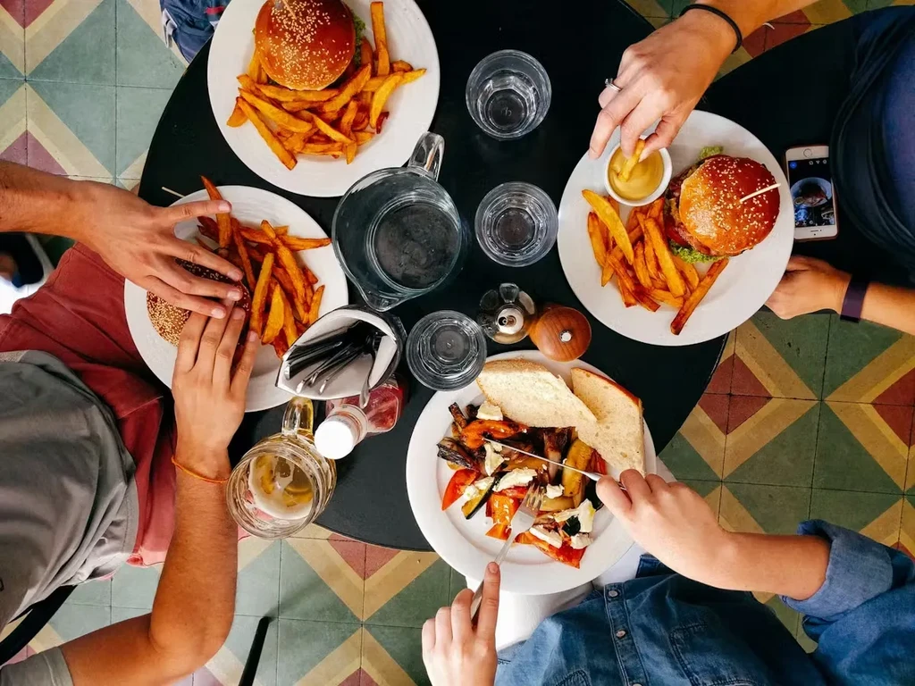 Hands eating burgers, fries, and vegetables from a dark table, top-down view.
