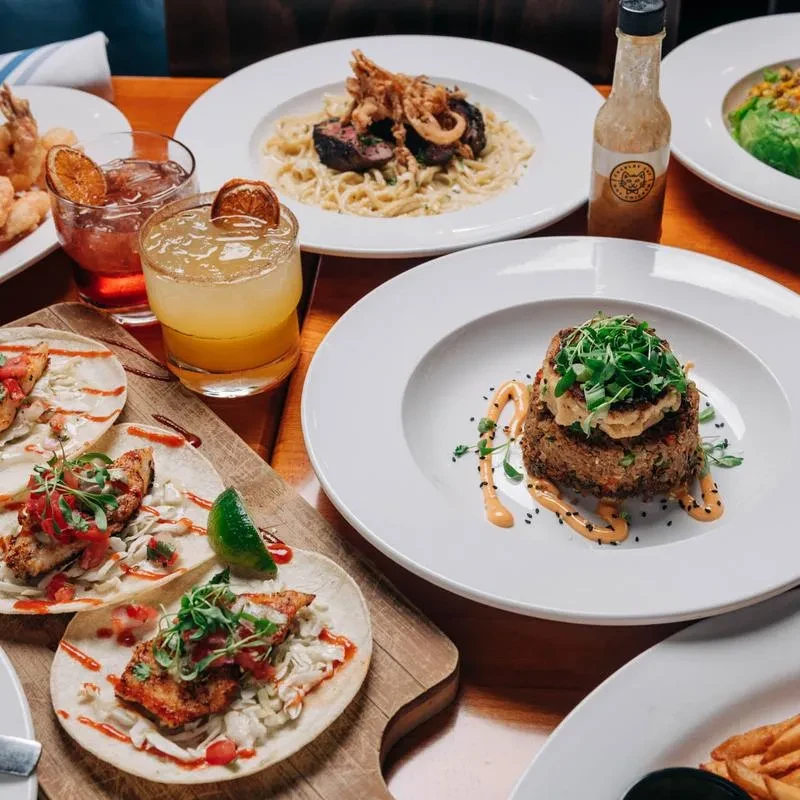 Three tacos, two cocktails, and diverse dishes on a wooden table.