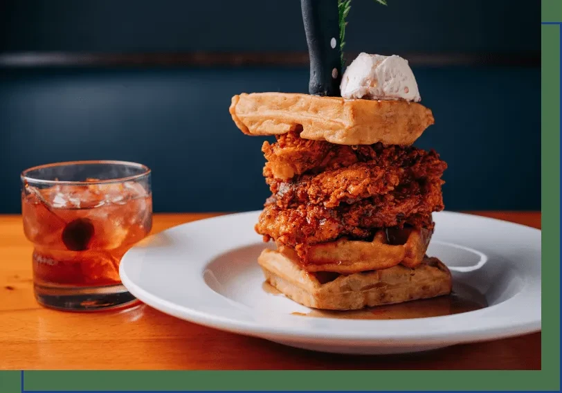 Stacked chicken and waffles with syrup, rosemary, and a cocktail.