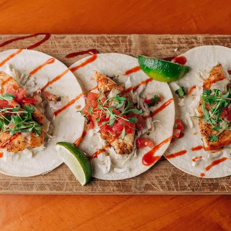 Three fish tacos on a wooden board with lime wedges and sauce.