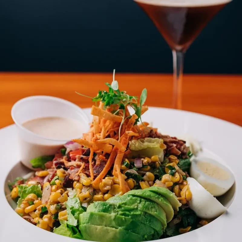 Colorful salad with corn, avocado, bacon, egg, dressing, and drink.