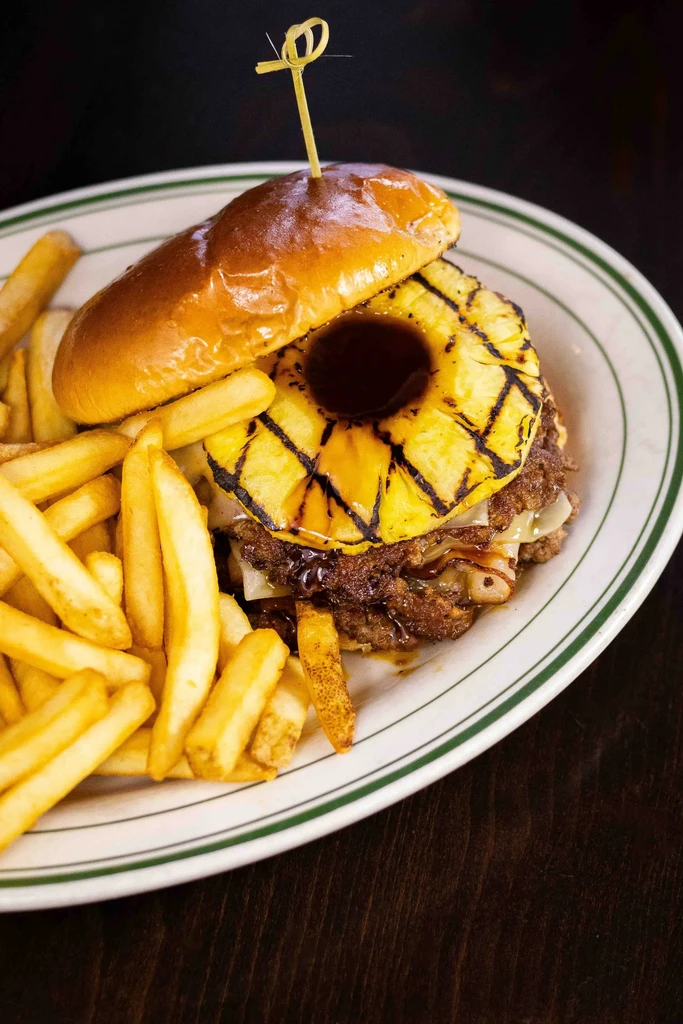 Gourmet burger with grilled pineapple, fries, on a striped white plate.