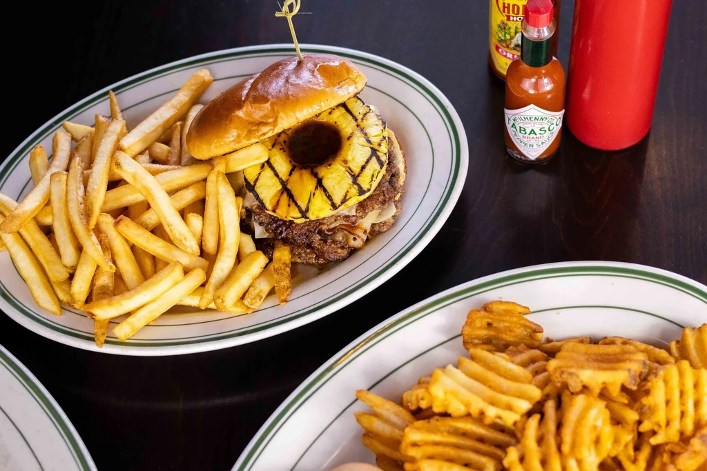 Overhead: burger with grilled pineapple, fries, and Tabasco on table.
