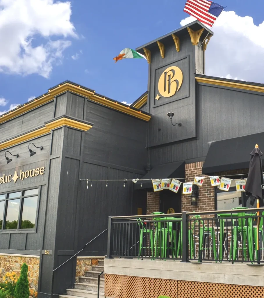 Exterior of a gold-trimmed public house with flags and a green patio.