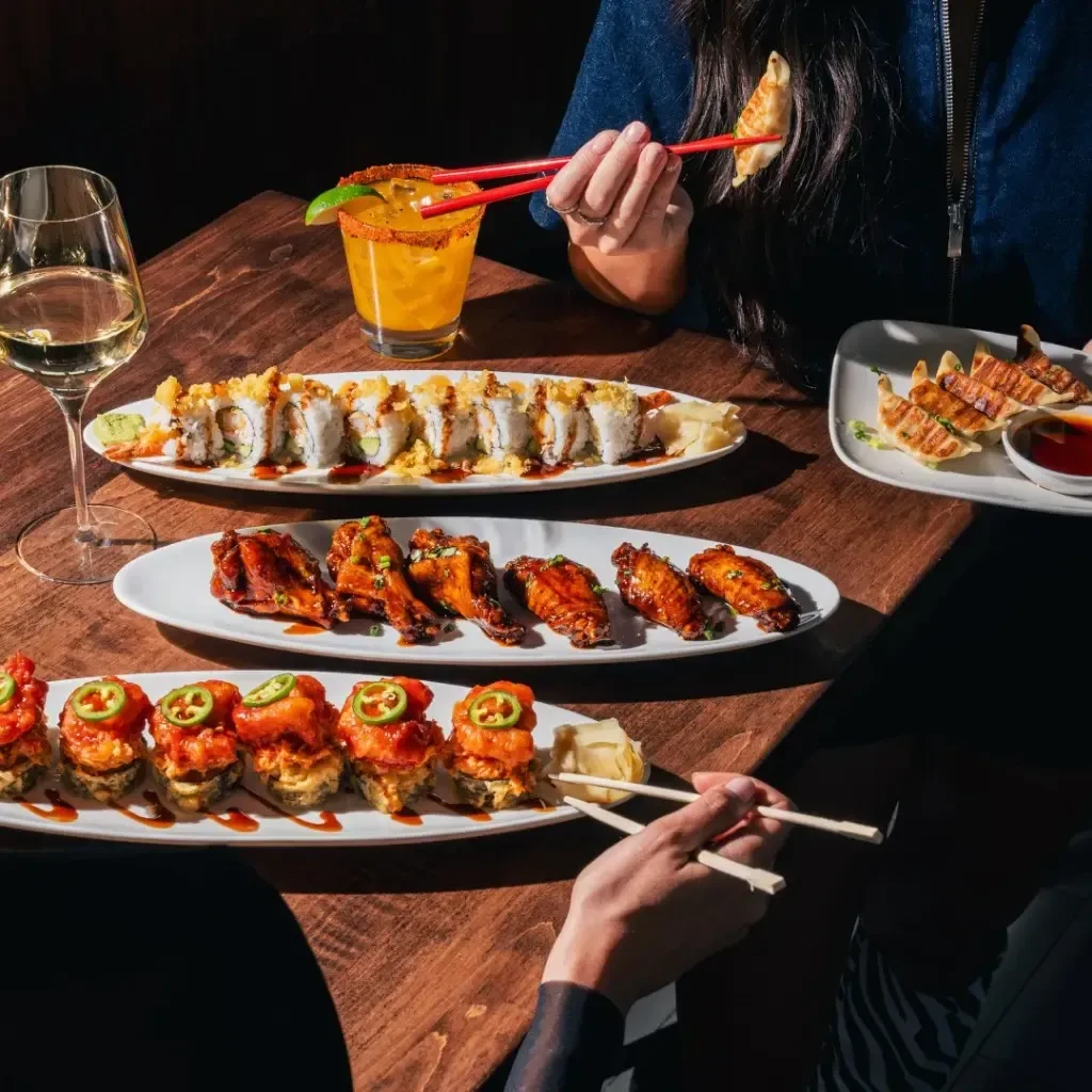 Overhead view: hands with chopsticks picking from sushi, dumplings, wings.