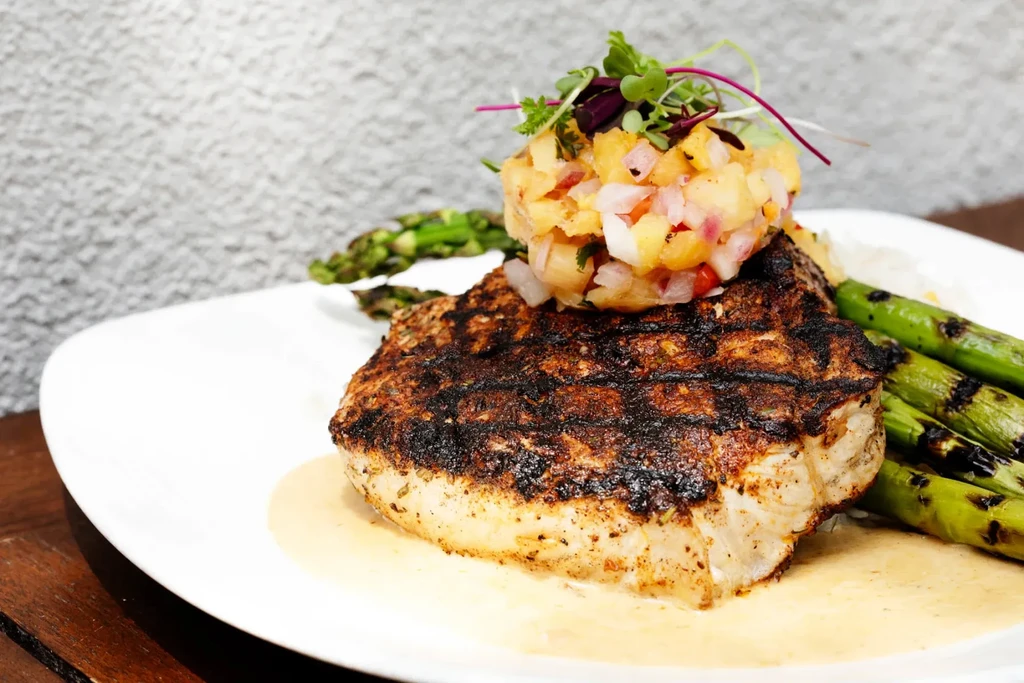 Grilled fish with fruit salsa, microgreens, and asparagus on a white plate.