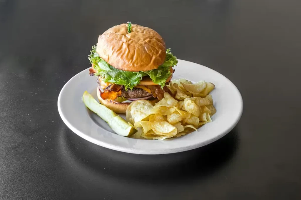 Bacon cheeseburger, golden chips, and pickle spear on a white plate.