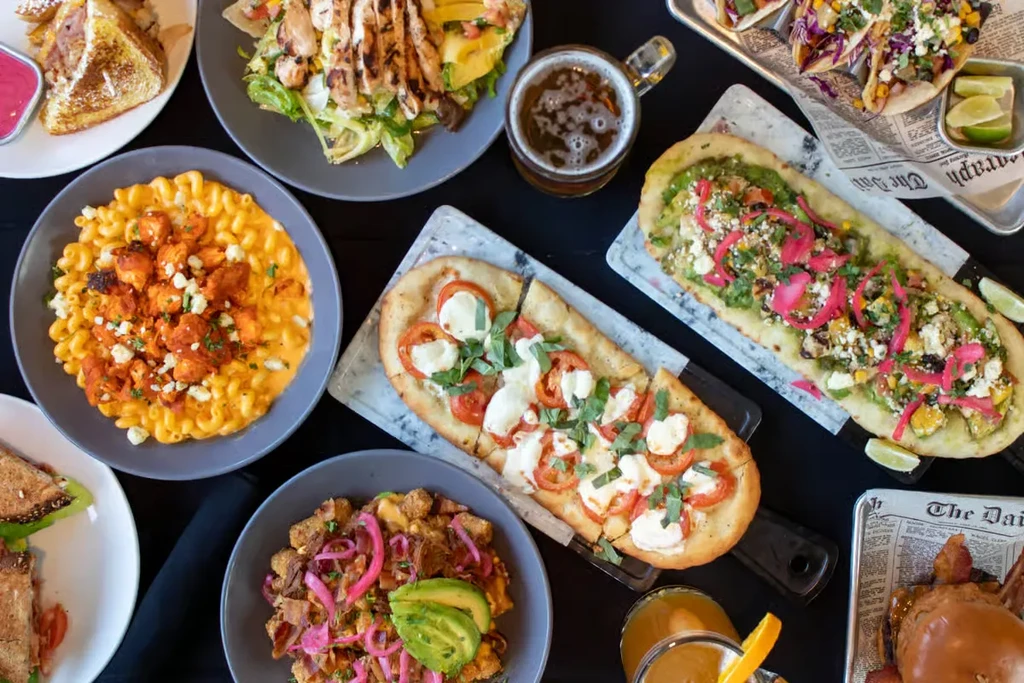 Overhead view of diverse restaurant dishes: flatbreads, tacos, salads, and drinks.