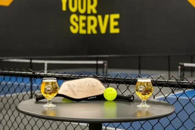 Pickleball paddle, ball, and beers on table with 'YOUR SERVE' sign.
