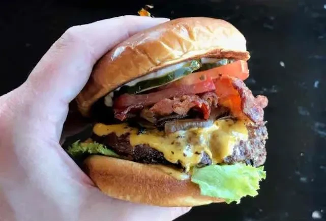 Hand holds a well-stacked burger with cheese, bacon, tomato, and lettuce.