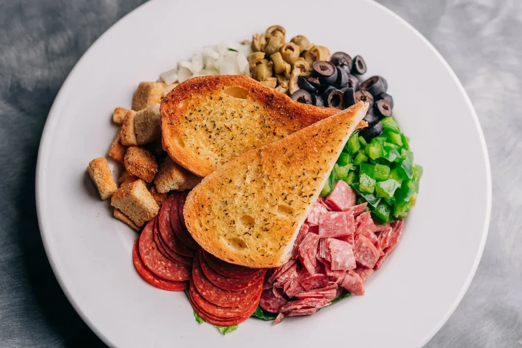 Overhead colorful salad with garlic bread, pepperoni, olives, peppers, and salami.