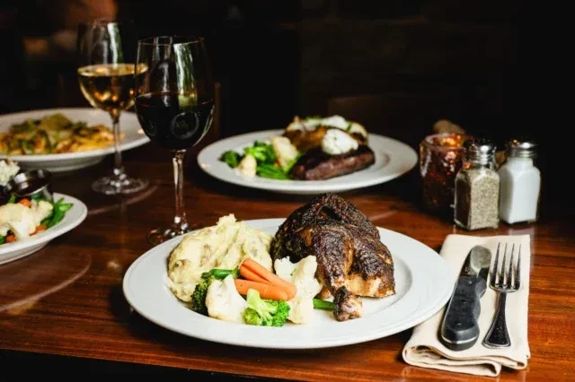 Restaurant table with roasted chicken, steak, vegetables, and wines.