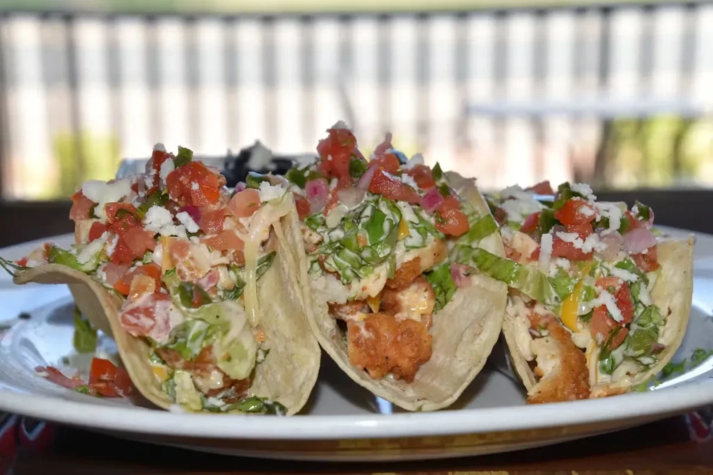 Three tacos with crispy fried protein, lettuce, tomato, onion, cheese.