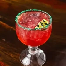Red cocktail in stemmed glass with green sugar rim and fruit wedge.