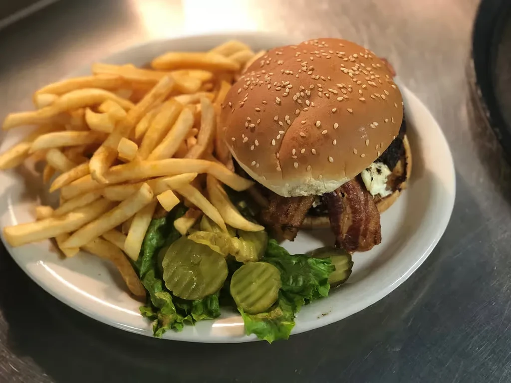 Large burger with bacon, fries, lettuce, and pickles on a white plate.