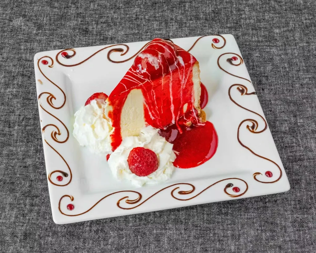 Cheesecake slice with strawberry sauce, white glaze, whipped cream, fresh strawberries.