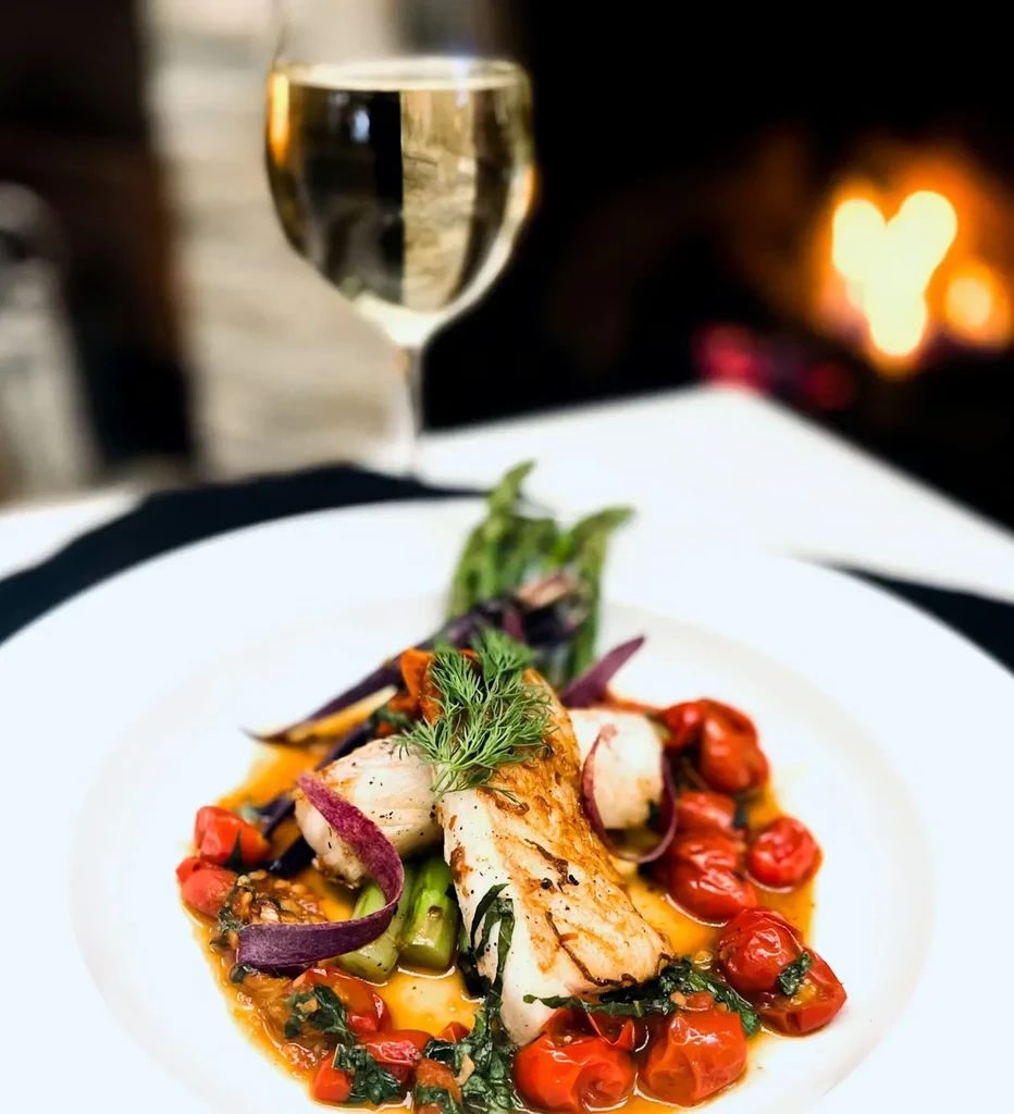 Plated fish/poultry, asparagus, tomatoes, radish, wine glass, fireplace.