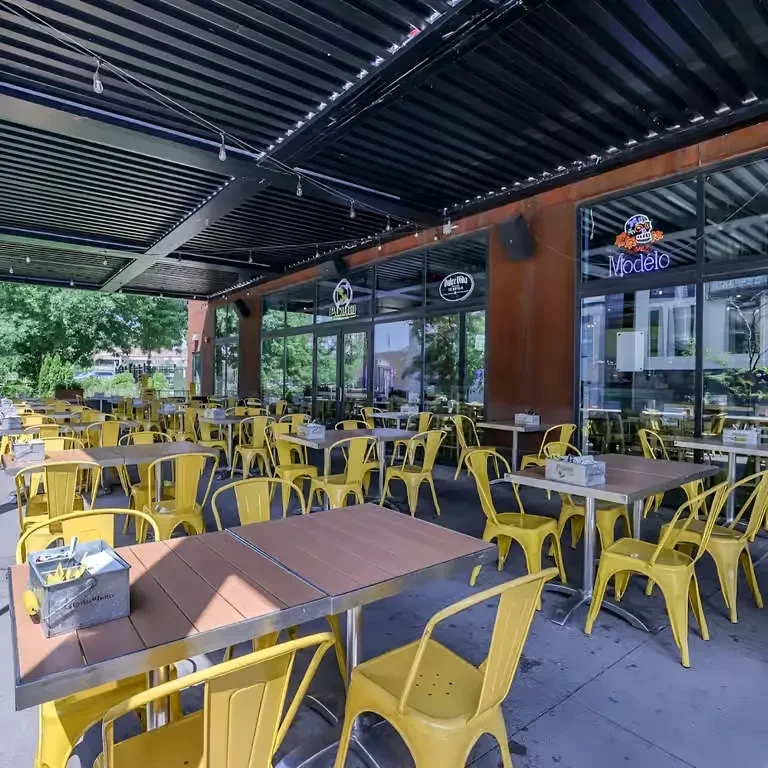 Modern covered patio with yellow chairs, wooden tables, Modelo neon sign.