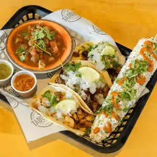 Assortment of Mexican tacos, elote, soup, and salsas in a basket.
