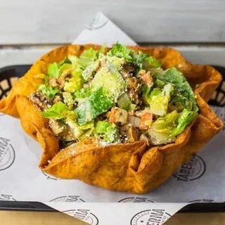 Caesar salad in a golden fried tortilla bowl on branded paper.