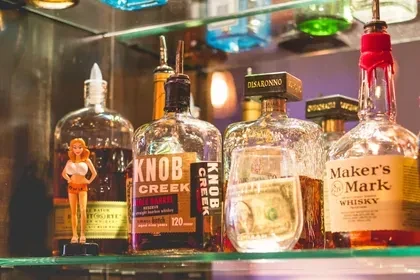 Liquor bottles on a bar shelf with a figurine and crumpled dollar.