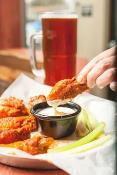 Hand dipping buffalo chicken wing into white sauce, with beer and celery.