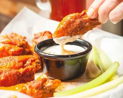 Hand dips orange Buffalo wing into creamy dip with celery.