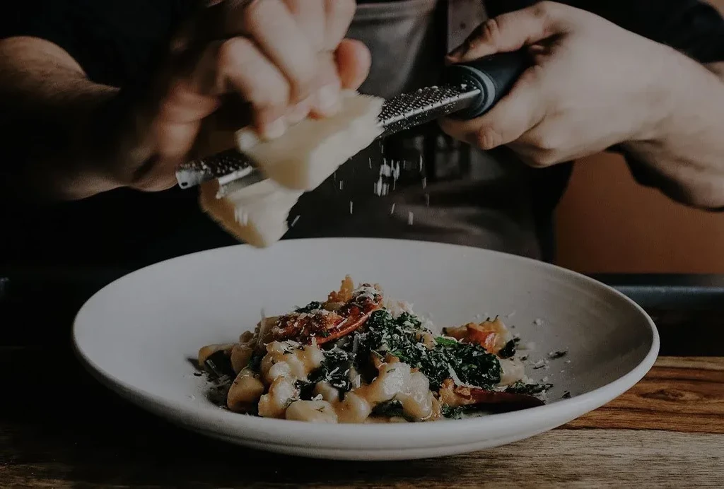 Hands grating cheese onto a bowl of gnocchi, spinach, and tomatoes.