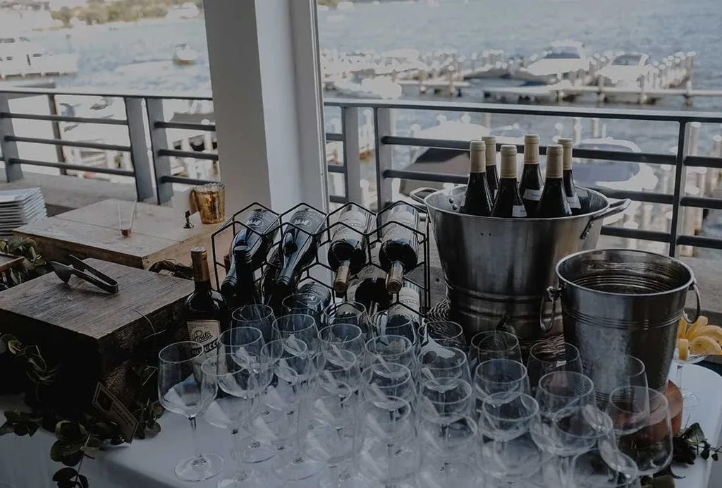Outdoor wine service table with glasses, bottles, and ice buckets overlooking water.