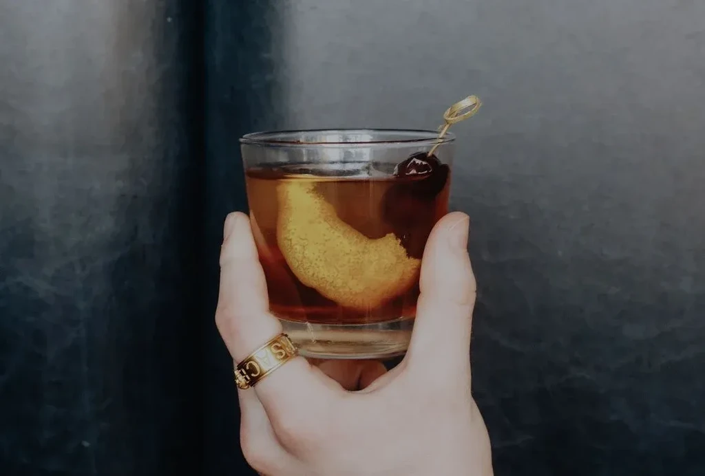 Hand holds amber cocktail with orange peel, cherry, and Versace ring.