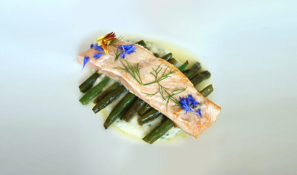 Plated salmon fillet with green beans, creamy sauce, dill, and flower petals.