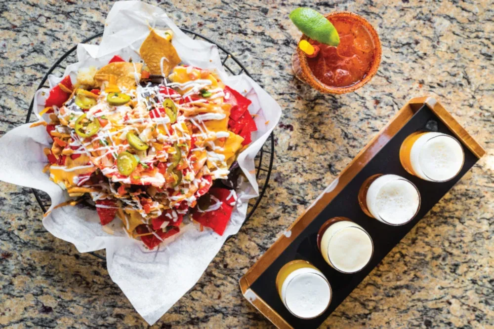 Overhead shot: loaded nachos, spicy cocktail, and beer flight.
