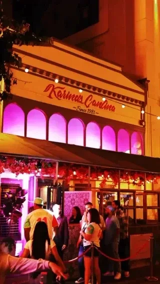 Karina's Cantina at night, yellow facade, magenta neon windows, queuing people.
