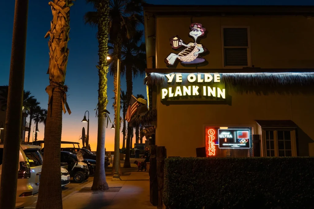 Illuminated beachside bar 'Ye Olde Plank Inn' with neon and palm trees.