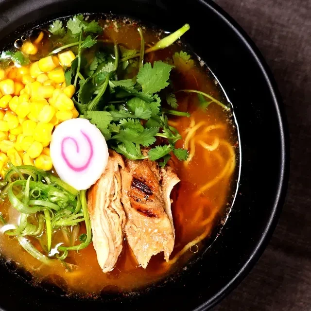 Steaming ramen bowl garnished with corn, chicken, narutomaki, cilantro.