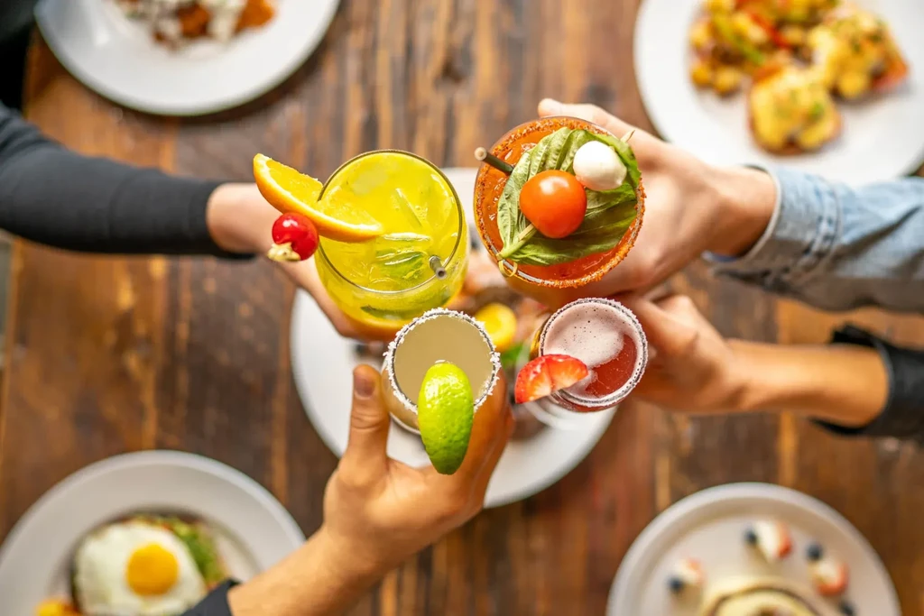 Four vibrant cocktails cheered by diverse hands at a lively meal.