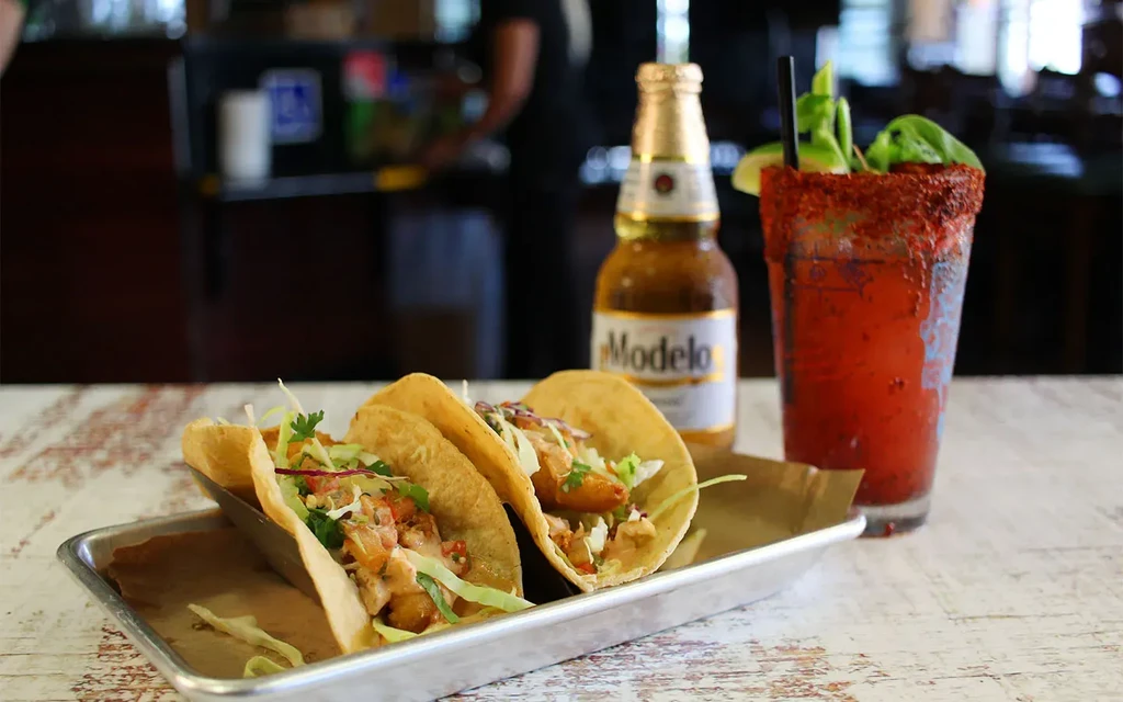 Three fish tacos, cold Modelo beer, and chili-rimmed drink on wooden table.