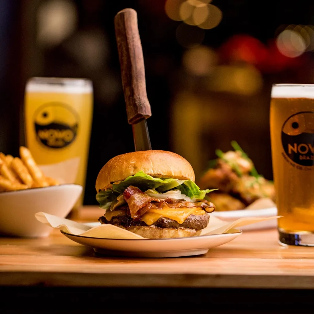 Bacon cheeseburger held by knife, with fries and two beers.