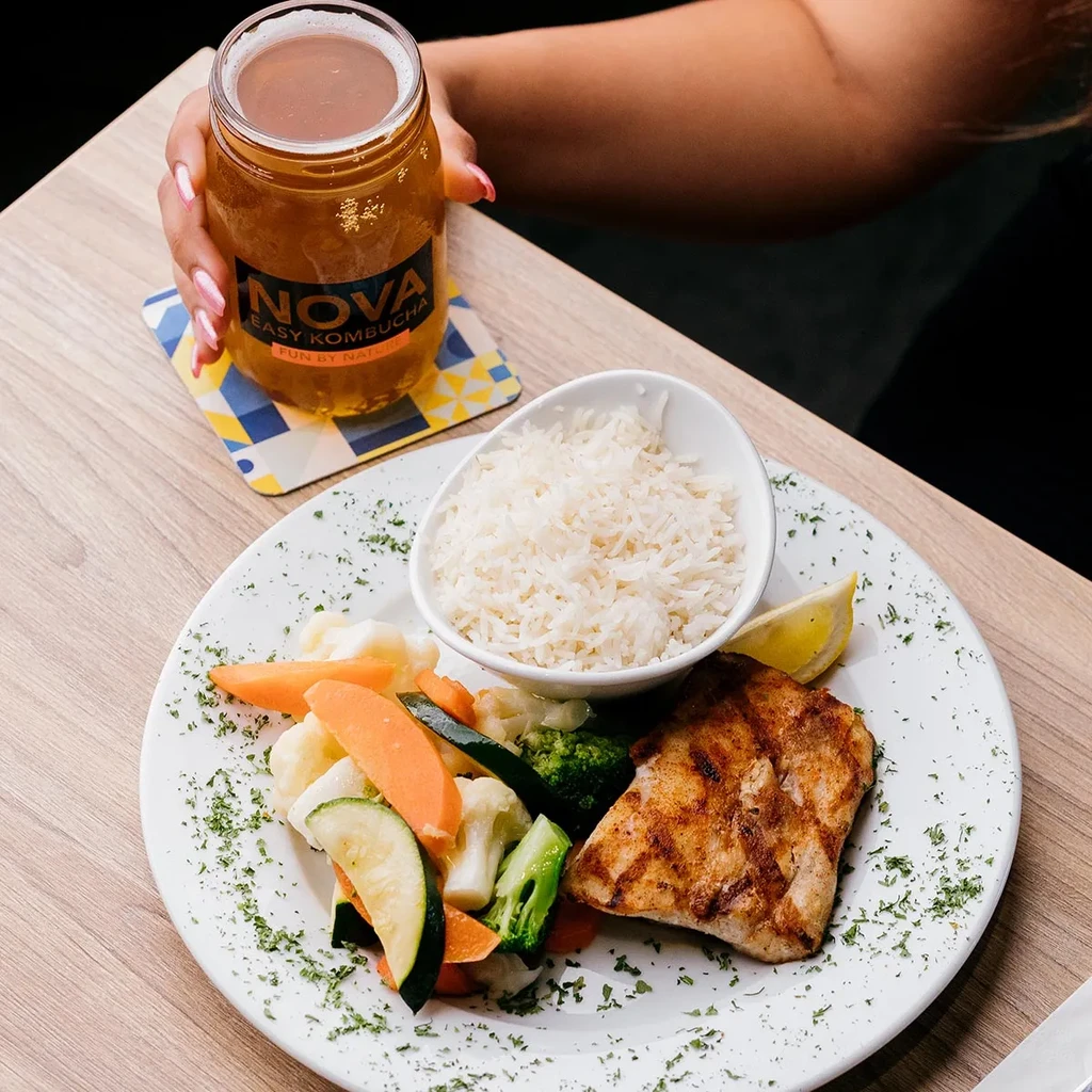 Grilled fish meal with vegetables, rice, and a hand holding kombucha.