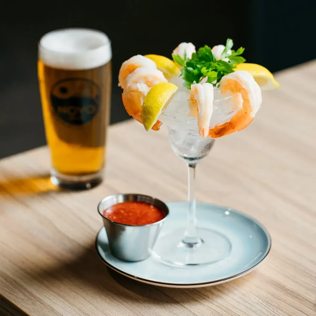 Shrimp cocktail, beer, and cocktail sauce on a light wooden table.