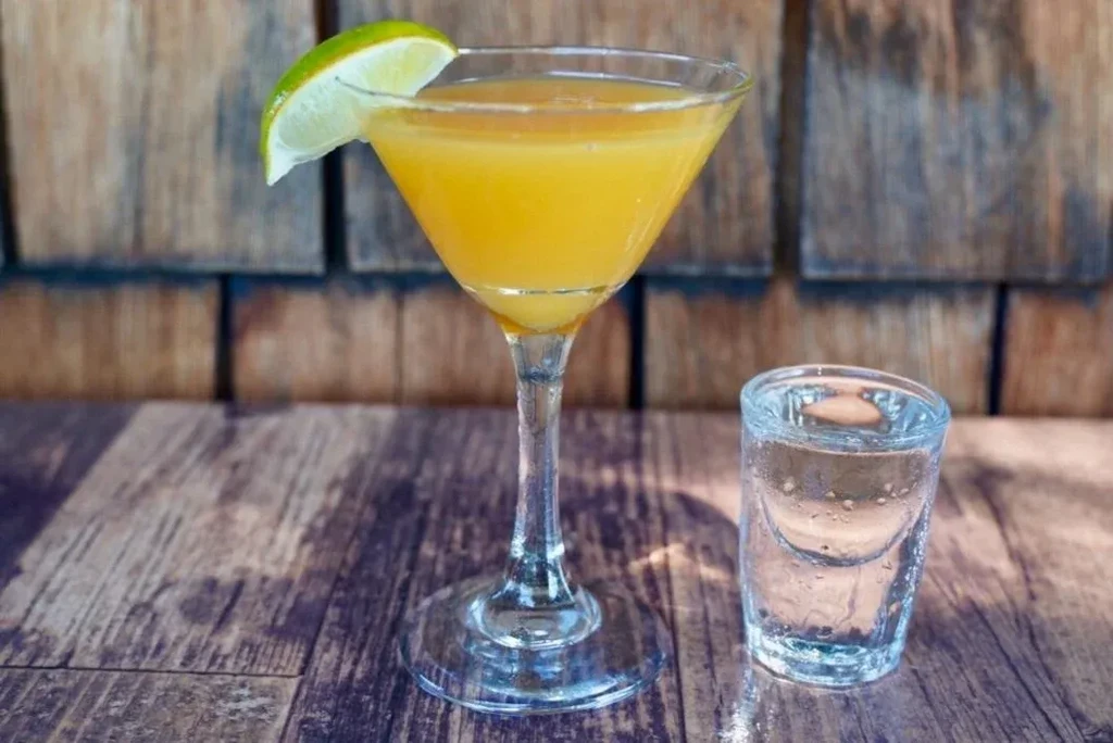 Orange martini with lime and clear shot glass on rustic wood.