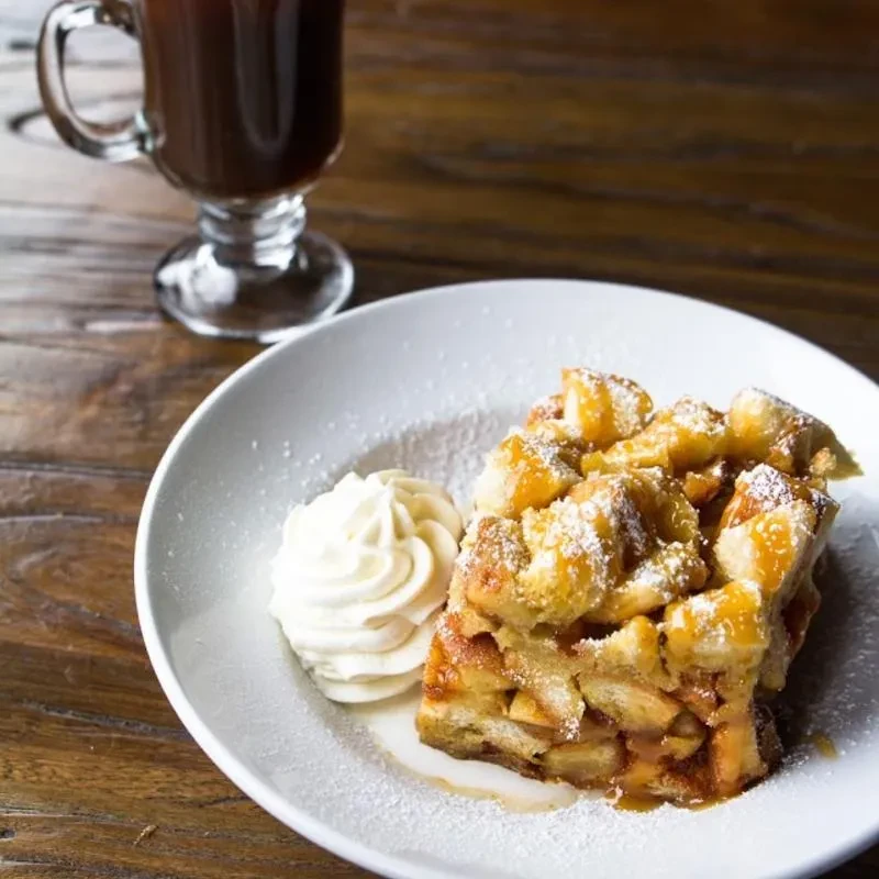 Golden-brown apple crisp or bread pudding, whipped cream, hot drink on wood.