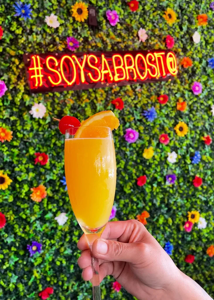 Hand holding mimosa with orange and cherry; neon sign, floral wall.
