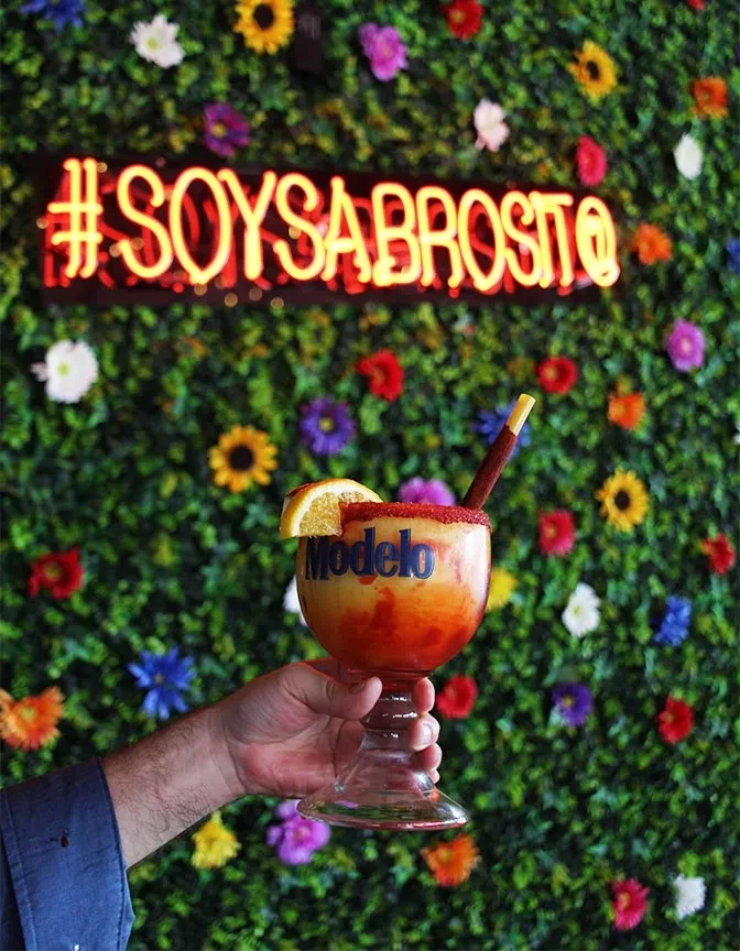 Hand holding Modelo cocktail with garnished rim against floral wall.