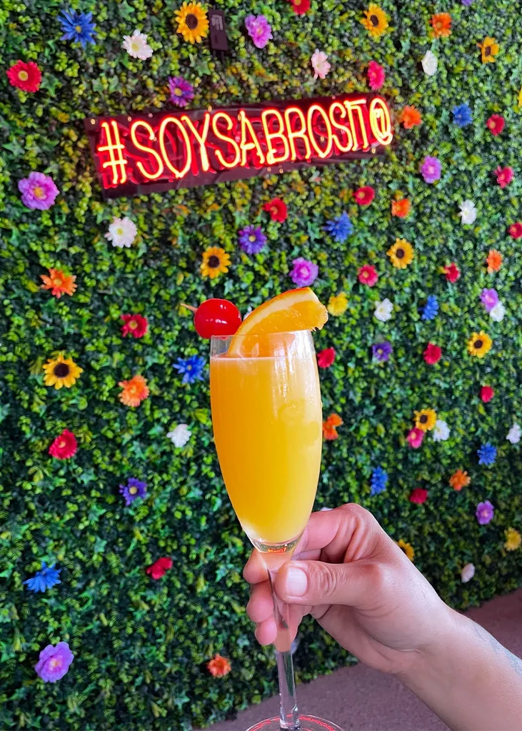 Orange drink in flute, hand, colorful flowers, and #SOYSABROSITO neon sign.