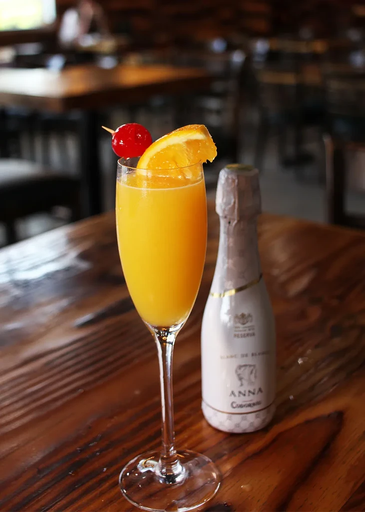 Mimosa cocktail, orange slice, cherry, and Cava bottle on wooden table.