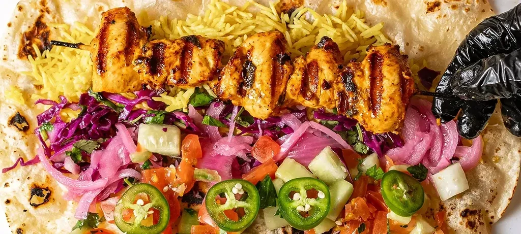 Gloved hand holds chicken skewer over open wrap, rice, vegetables.
