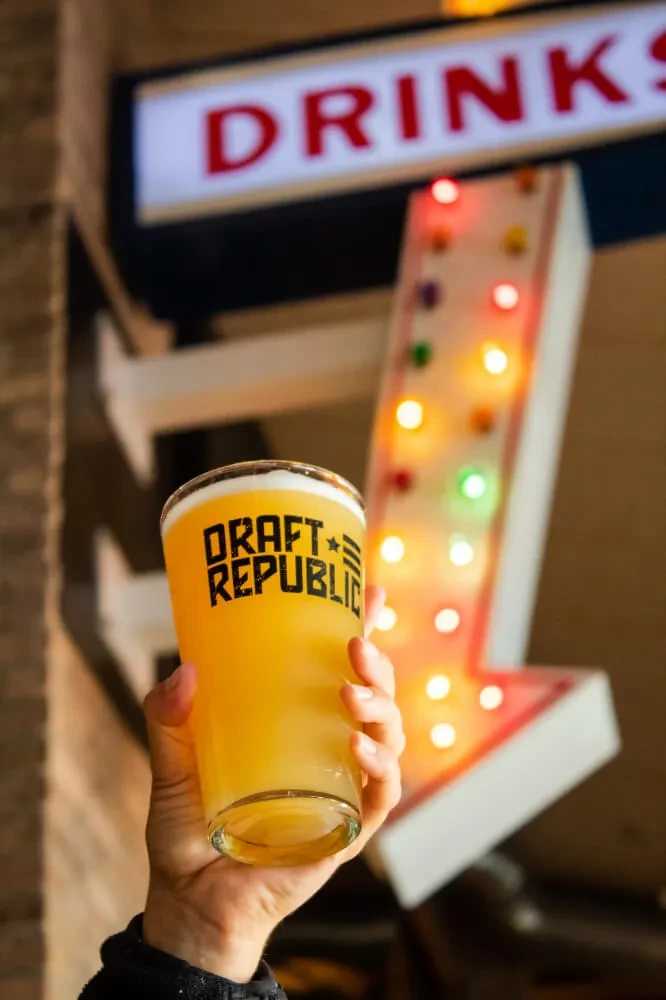 Hand holding Draft Republic beer glass; blurred illuminated bar signs.