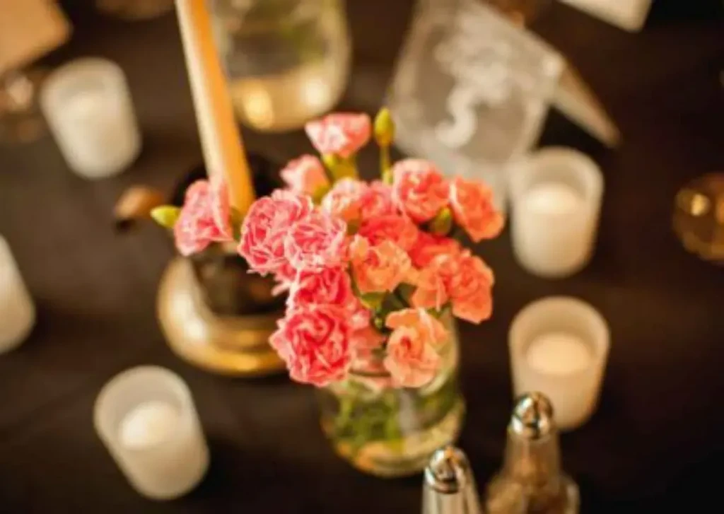 Coral carnations, taper candle, and lit votives on a dark dining table.