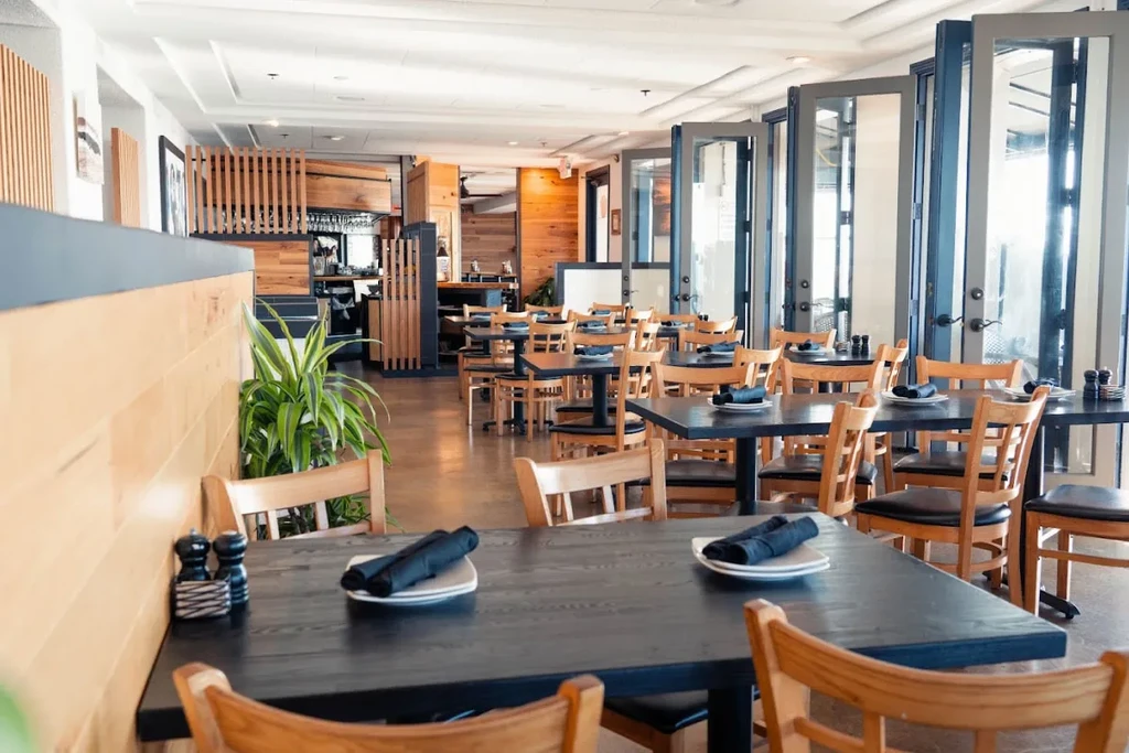 Modern restaurant interior with dark tables, light wood chairs, and glass doors.